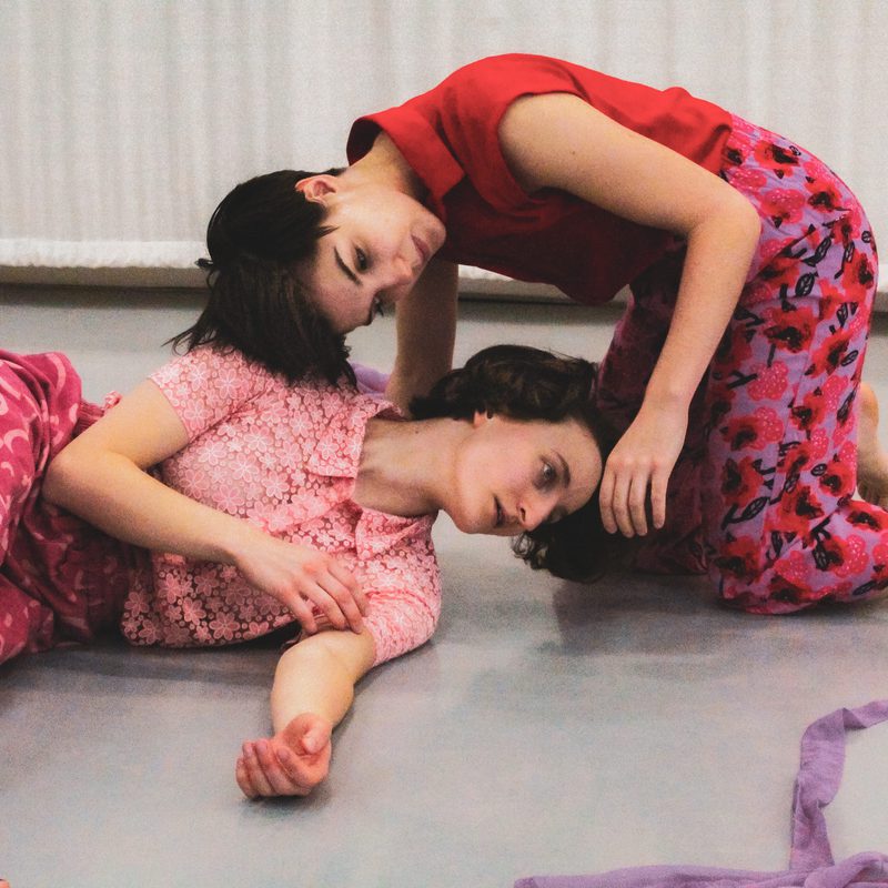 The image features two white female dancers with short dark hair. The floor and the background are both white. They are both wearing pink patterned trousers and short-sleeved blouses. One of the dancers is lying on the floor, while the other dancer is kneeling and bending over them with their head gently touching the other dancer’s shoulder.