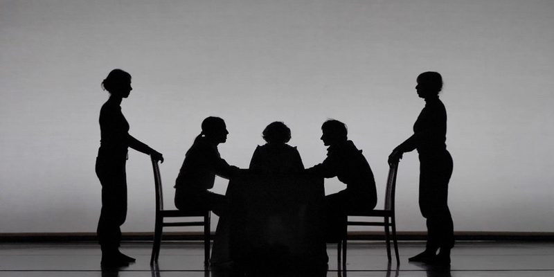 Black and white image of five bodies onstage. Three are sitting on a table facing each other. Two are standing behind the chairs that two of the bodies are sitting on.