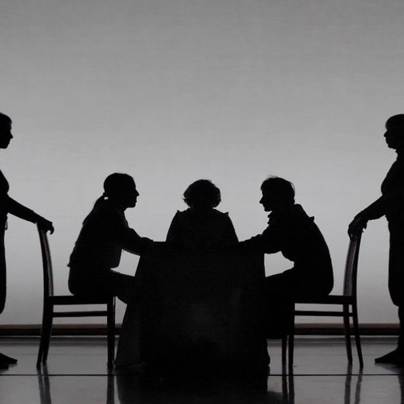 Black and white image of five bodies onstage. Three are sitting on a table facing each other. Two are standing behind the chairs that two of the bodies are sitting on.