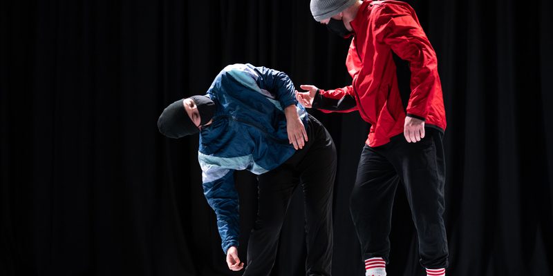 Two dancers in the image, one is leaning down to the right whilst the other looks on. One wears a blue jacket and a black hat with a mask, the other a red jacket, grey hat and a mask.