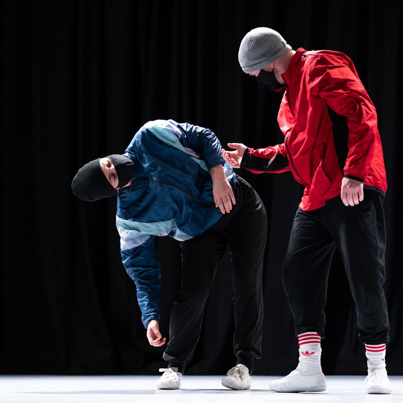 Two dancers in the image, one is leaning down to the right whilst the other looks on. One wears a blue jacket and a black hat with a mask, the other a red jacket, grey hat and a mask.