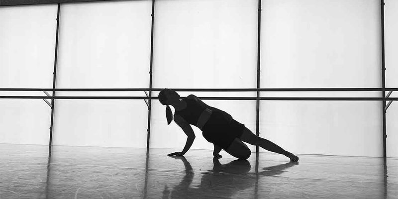 Black and white image of a dancer in the studio. A barre is in the background running through the image. There are also windows. The dancer kneels down on one leg with the other leg out. They also have their hand on the floor which, along with their knee, they are using to support themselves.