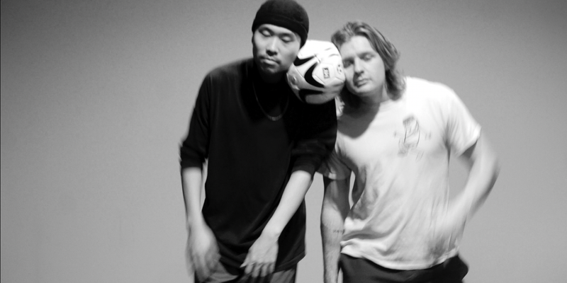 Image of two dancers. They hold a football between their heads. One dancer is wearing black and has a black hat on. The other is wearing a white graphic t-shirt.