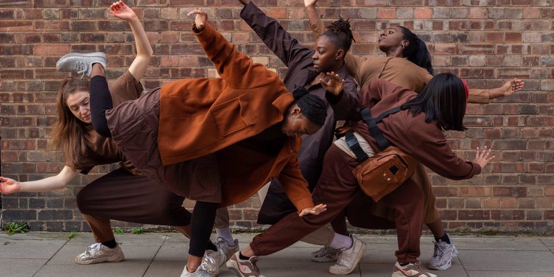 Image of five dancers all in a group huddled together. They make different shapes with their arms and bodies.