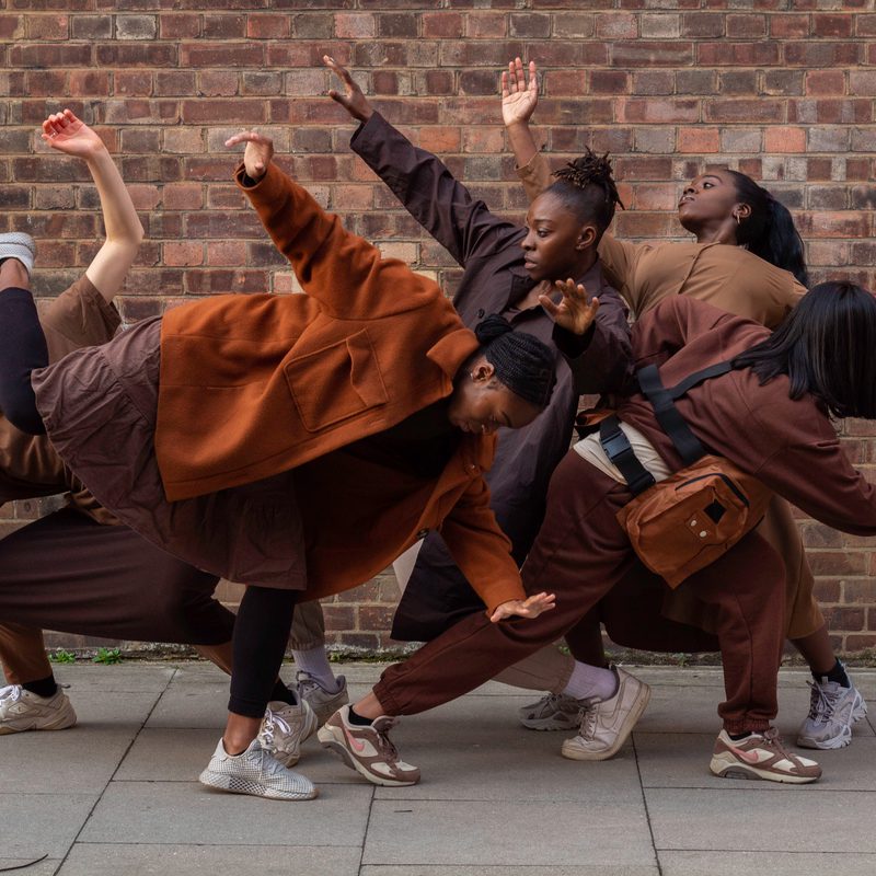 Image of five dancers all in a group huddled together. They make different shapes with their arms and bodies.