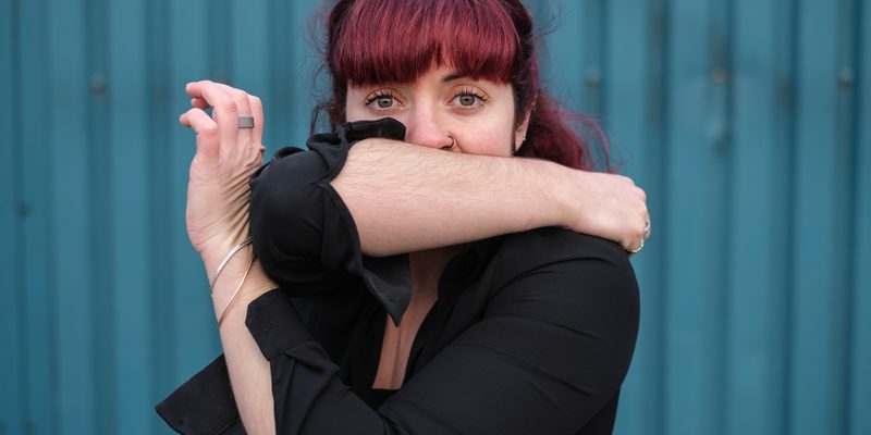 Image of a body in the frame with pink hair. They cover their mouth with their right arm and cross their left arm across their chest so their elbow and left hand connect.