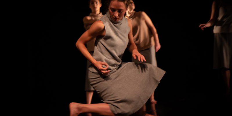 Image of a dancer dressed in a grey top and grey dress kneeing down, turning slightly to the right so their right knee is hovering off the floor. Their arms are bent slightly so their hand is by their hip.