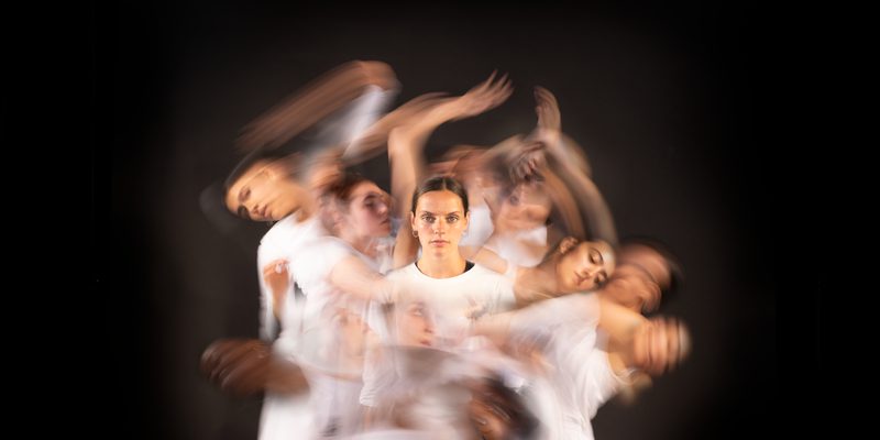 A tight knit group of dancers swirl around a central figure, creating a nebulous swirl of movement centred around a single dancer.