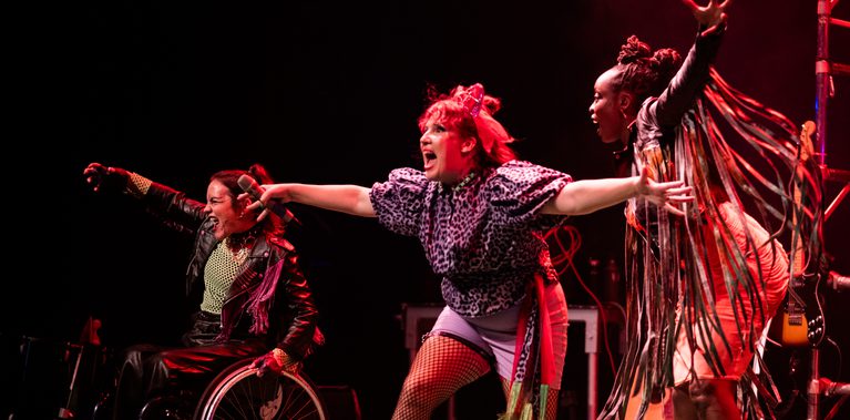 3 performers on stage, dressed in punky costumes of leather, tassels and patterns have their arms and mouths wide as they move energetically across the stage in different directions.