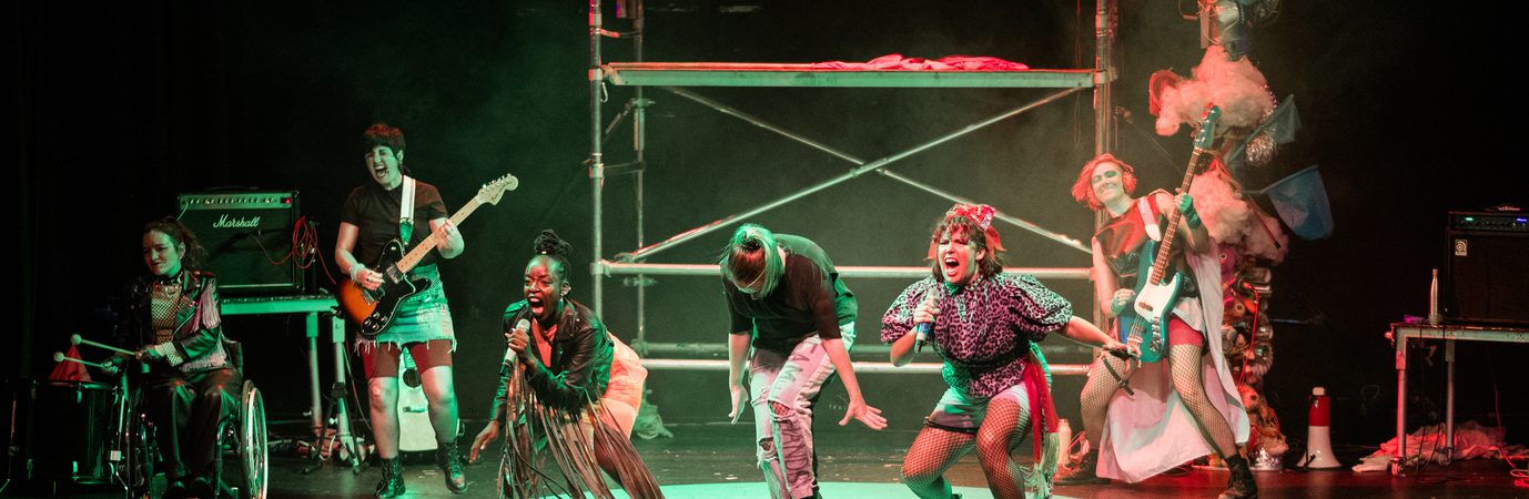 On stage, the band all dressed in punk clothing are rocking out. The full stage which has the backdrop of a scaff tower which red and green light reflects off. The 6 performers are singing, playing bass and guitar and on the drums.