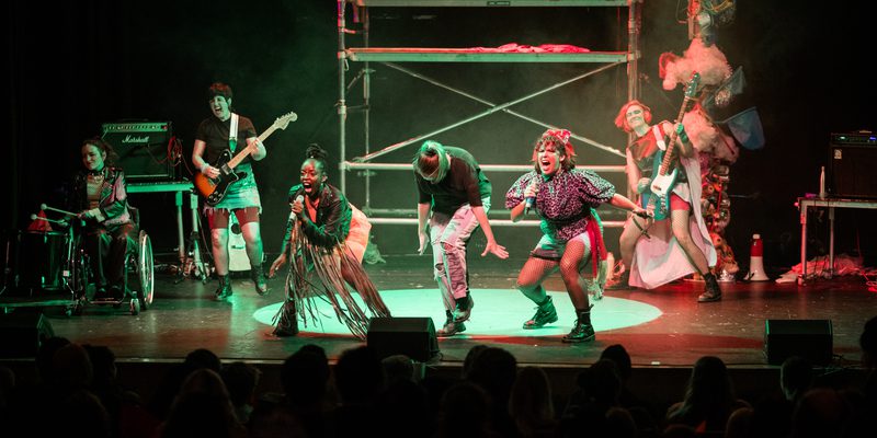 On stage, the band all dressed in punk clothing are rocking out. The full stage which has the backdrop of a scaff tower which red and green light reflects off. The 6 performers are singing, playing bass and guitar and on the drums.