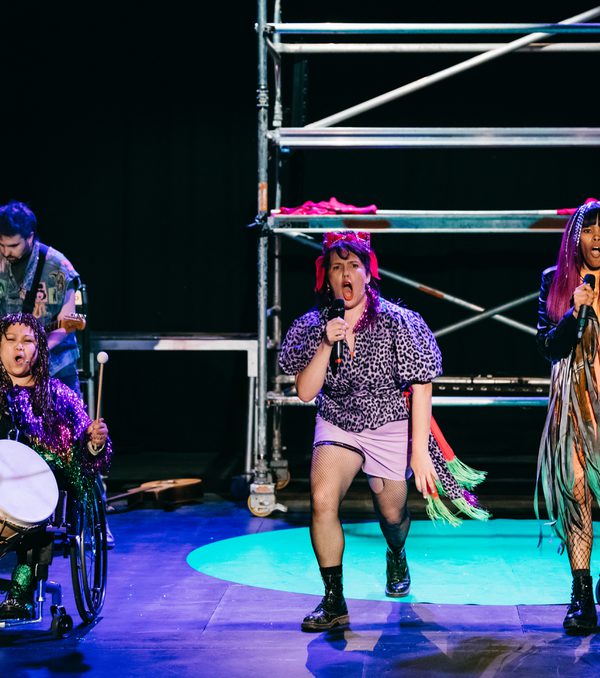 3 band members stand at the front of the stage singing. They are dressed like a punk band, some singing, 1 playing a drum.