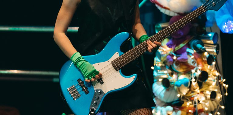 Rake is on stage wearing a punk outfit and headphones, they are playing a bass guitar.