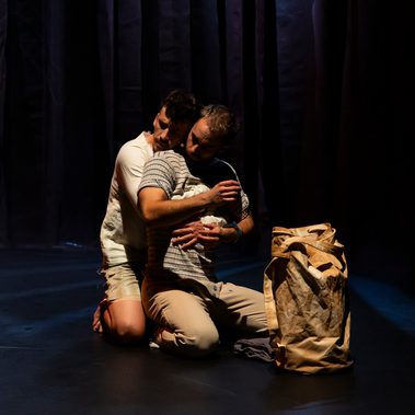 A tender scene. Two male presenting dancers are kneeling on the floor, one in front of the other, arms wrapped tightly around each other’s chest. It is Mariposa and the sailor Preston. Is this the start of a passionate love affair? A duffle bag is positioned in the foreground of the shot.