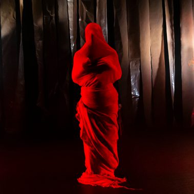 A solo figure wrapped mummy-like from head to toe in bright red coloured cloth stands alone on stage. Both hands are held closely to the chest, the head pointing slightly up and the overall posture conveying the anguish and pleading of someone who has been crushed emotionally.