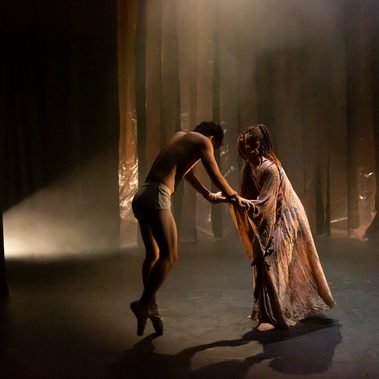 A tall male presenting dancer, dressed only in white shorts is standing on pointe, leaning slightly forward, head bent with his arms outstretched gives the impression of being weak and fragile. He’s leaning in towards a female presenting dancer who is holding both his hands in support and looking intently into his face.