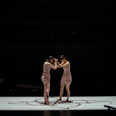 A wide image showing a pitch-black stage, and a white floor. Two women are facing each other, their hands locked together and their heads down. They wear skin-coloured shifts, and their feet trace inky-black circles on the white floor.