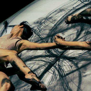 From above we see a white floor, with expressive inky black marks across it, and two women grasping each other by the hand with their limbs spread around.