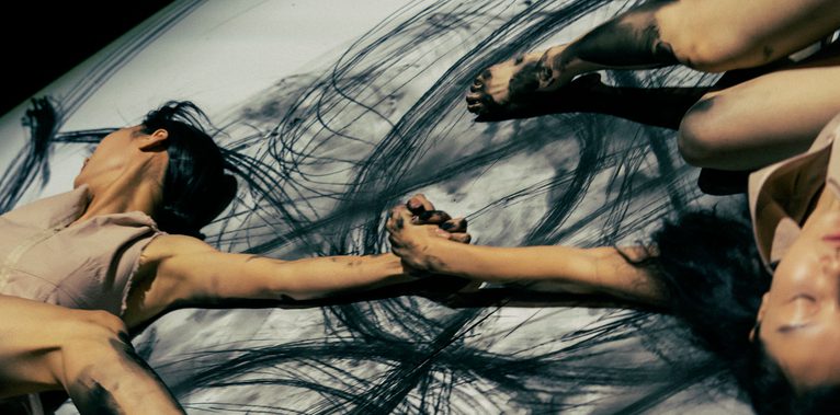 From above we see a white floor, with expressive inky black marks across it, and two women grasping each other by the hand with their limbs spread around.