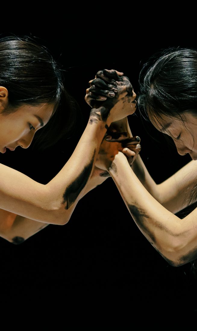 Two women face each other, with their hands locked into each other's, and their heads down, against a dark black stage. Their arms and hands are covered in a black inky substance.