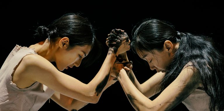 Two women face each other, with their hands locked into each other's, and their heads down, against a dark black stage. Their arms and hands are covered in a black inky substance.