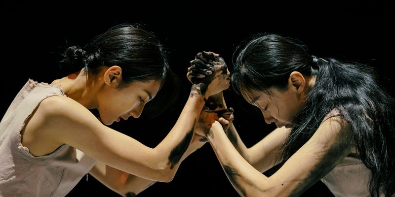 Two women face each other, with their hands locked into each other's, and their heads down, against a dark black stage. Their arms and hands are covered in a black inky substance.