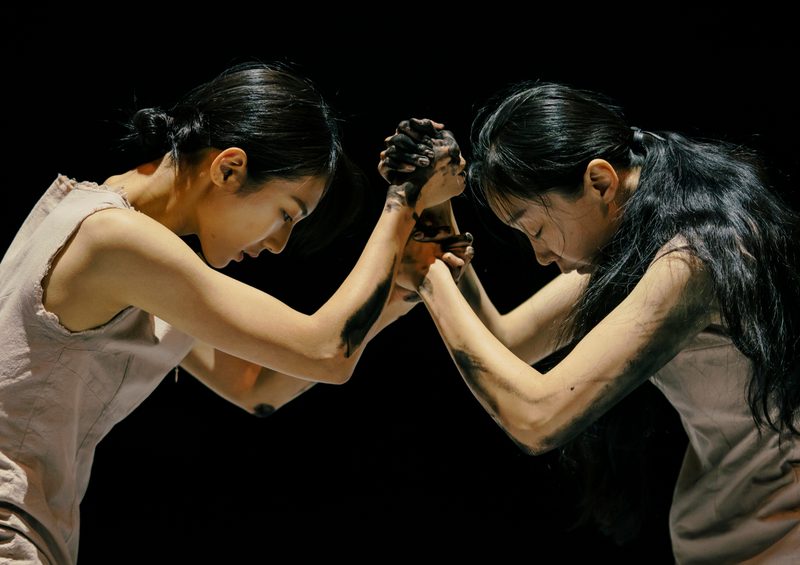 Two women face each other, with their hands locked into each other's, and their heads down, against a dark black stage. Their arms and hands are covered in a black inky substance.