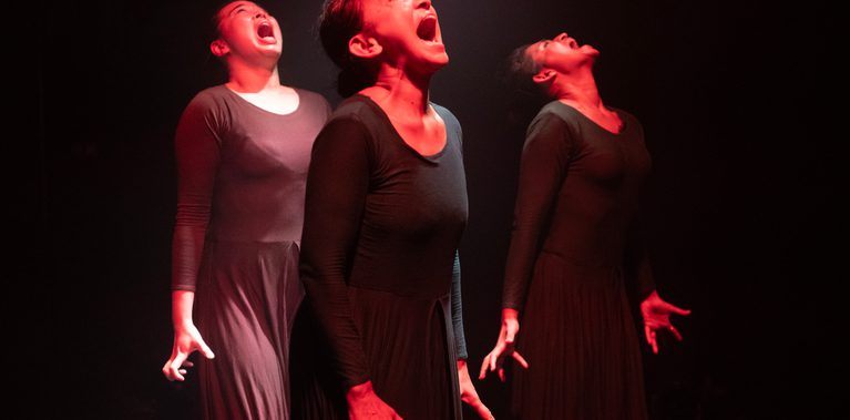 Three female dancers bathed in red stage light - standing straight and silently screaming with their mouths wide open and heads tilted back and wearing an agonised expression and displaying their trauma as a result of domestic abuse.