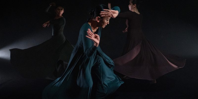 Three female dancers positioned in a triangle shape. One of the two dancers at the back is charging towards the other in a fighting mode to hurt her while the dancer in front tries to stay out of their way feeling vulnerable knowing they will come after her next. The caption also displays the wild movement of their swirling long dresses.