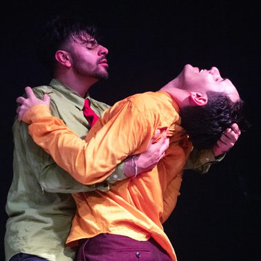 A male dancer in a light green shirt embracing another male dancer in a yellow short - pulling the latter’s head back by his hair. Their eyes are closed, expressing passion and desire of each other – confident in expressing their relationship.