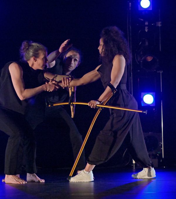 Three dancers, Annie Hanauer, Giuseppe Comuniello, and Laila White, all dressed in black stand in a group facing one another on a stage with a a black background and blue lighting behind them. Annie, to the left, holds with both hands the right wrist of Laila to the right, who also holds two gold crutches. Giuseppe, between them, points above the clasped hands with his right hand.