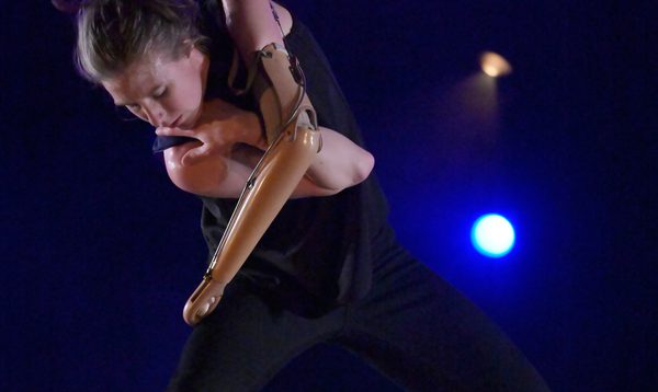Annie Hanauer wearing black stands on a stage with a black background and blue lights behind. She has a wide stance and leans forward with a prosthetic left arm crossed over a bent right arm.