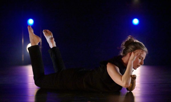Annie Hanauer, dressed in black trousers and sleeveless top, lays on her stomach on a dark stage with blue lights behind. Her feet point upwards and her upper body is raised with her right arm near her ear.