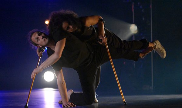 Annie Hanauer, Giuseppe Comuniello, Laila White and Deborah Lennie, all dressed in black on a dark stage with blue and white lights behind. They are gathered into a tight group with Deborah and Laila facing away and only Giuseppe’s feet visible. Annie’s weight is supported by the group and she faces the camera with her feet suspended off the ground.