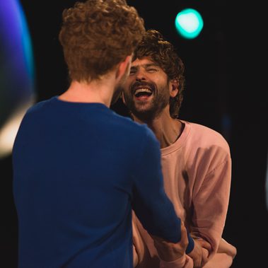A close up shot of three male performers, two caught in a happy embrace of the arms with wide smiling expressions and one overlooking the interaction from a distance. Blurred in the background is a green balloon hanging from the ceiling.