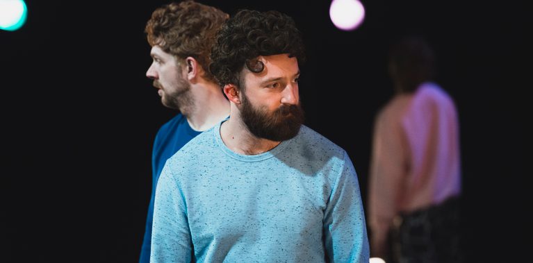 A close up shot of three male performers, two standing side by side yet facing looking outwards from one another and one facing away from us looking to the ceiling from a blurry distance.