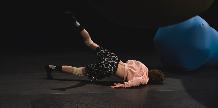 n image with a black background and black stage floor containing one blue balloon on the floor and one green balloon hanging from the ceiling. You can see a male performer in the centre on the floor, lying on his side with his back facing us. He has his left leg stretched in the air and is bearing weight on his shoulder.