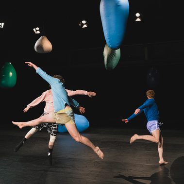 An image with a black background and black stage floor containing multiple large balloons of various colours (white, green, blue) with a string of stage lights surrounding the perimeter of the ceiling. You can see three male performers mid-air jumping with legs spread in a circle formation. You can not see any of their faces as they are either blocked by one another or facing away from us.
