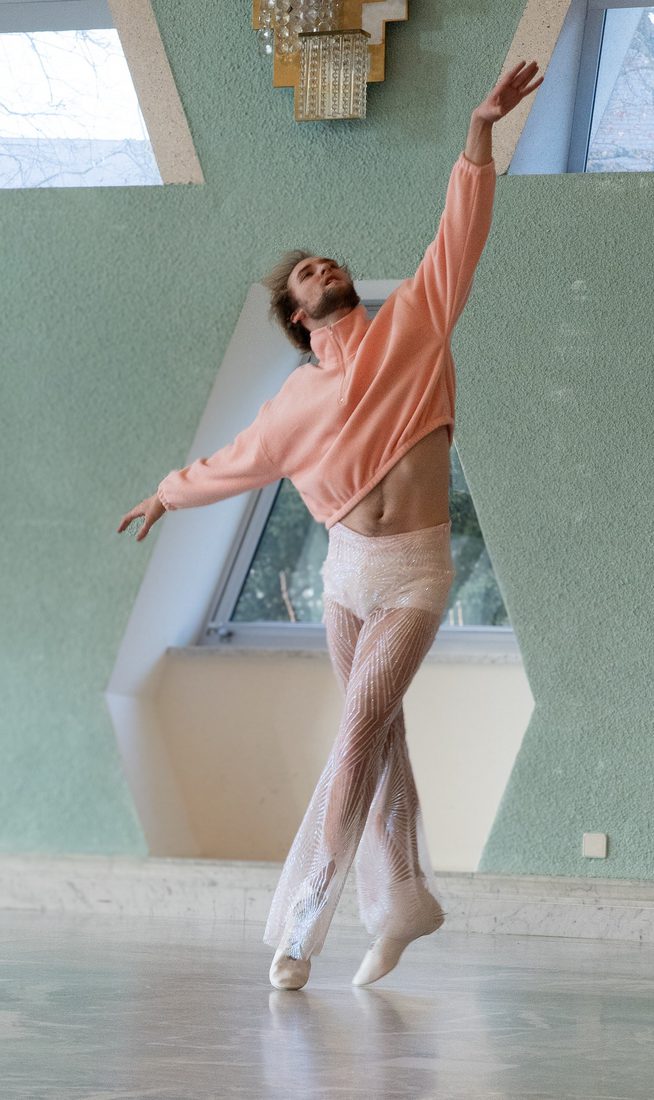 A contemporary dancer in a pink cropped sweater and sheer, sparkling trousers balances on pointe in a sunlit studio with mint green walls and geometric skylight windows.