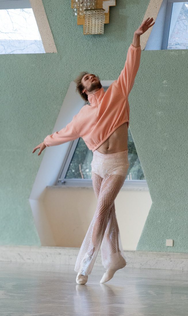A contemporary dancer in a pink cropped sweater and sheer, sparkling trousers balances on pointe in a sunlit studio with mint green walls and geometric skylight windows.