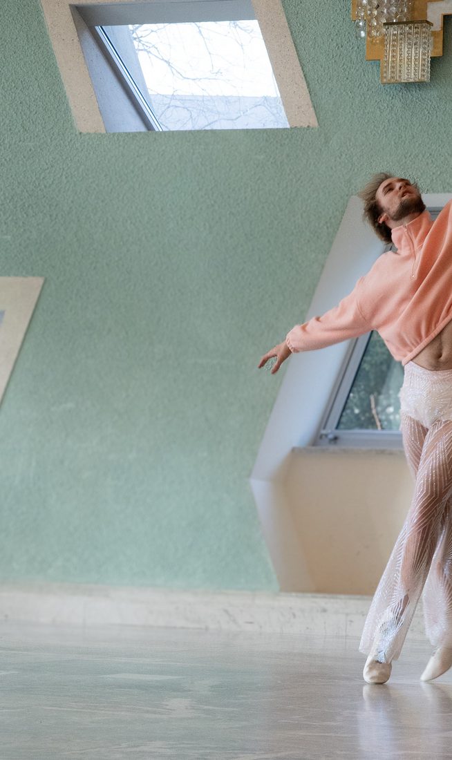 A contemporary dancer in a pink cropped sweater and sheer, sparkling trousers balances on pointe in a sunlit studio with mint green walls and geometric skylight windows.