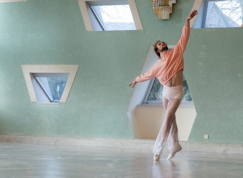 A contemporary dancer in a pink cropped sweater and sheer, sparkling trousers balances on pointe in a sunlit studio with mint green walls and geometric skylight windows.