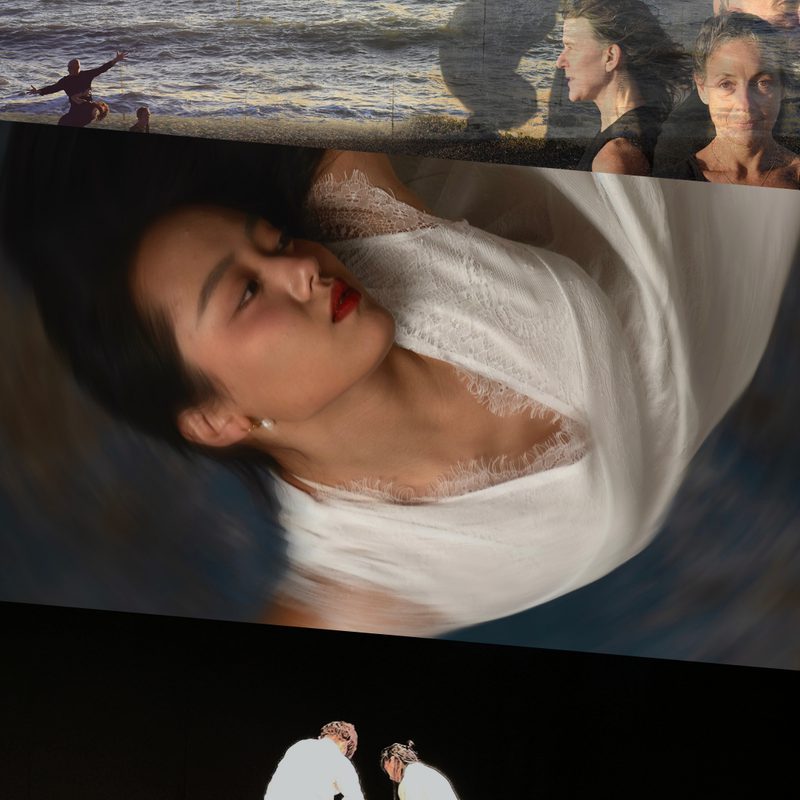 This image is split into three parts. On the top, a layered image featuring a sea with two distant figures standing before it, a light brick wall casting shadows of three posing figures, and three people shown from the shoulders up, one facing forward, with two behind her in profile facing right and left. In the centre, blurred image of a person with long black hair, seen upside down, wearing a long white dress, leaning sideways with arms raised to shoulder level and elbows bent at right angles. On the bottom, three people, all wearing white t-shirts and black trousers, are in an all-black theatre setting, with two standing back to the audience, lifting the legs of a third person lying on their stomach, looking forward and reaching ahead.