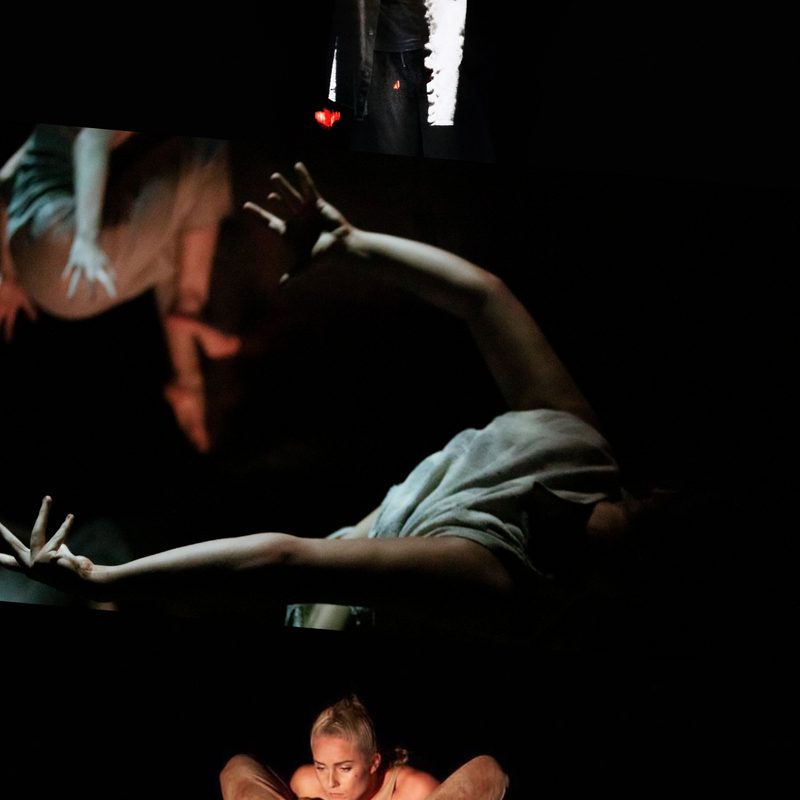 This image is divided into three parts. On the top, a person dressed in white stands in a dim spotlight against a dark background, holding their hands raised to chest level in a gesture, with their face hidden in shadow. In the centre, close-up of a person’s arms with fingers spread, reaching behind their back while lying face down in a lightly lit dark setting. On the bottom, two figures are illuminated by yellow light as one person squats holding another who is upside down, leaning backward and wrapping their thighs around the squatting person.