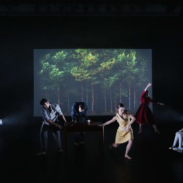 Six performers in motion on a dark stage, with the stage walls visible. Behind them is a projection of trees at night