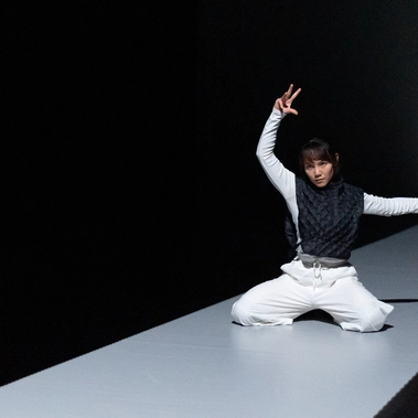 A dancer kneels on a narrow, illuminated stage, wearing white pants and a dark textured vest, striking a dramatic pose with one arm raised and the other extended outward.