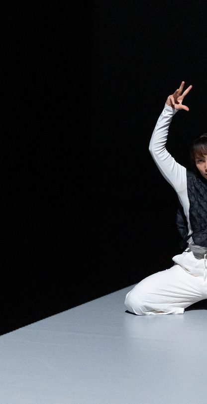 A dancer kneels on a narrow, illuminated stage, wearing white pants and a dark textured vest, striking a dramatic pose with one arm raised and the other extended outward.