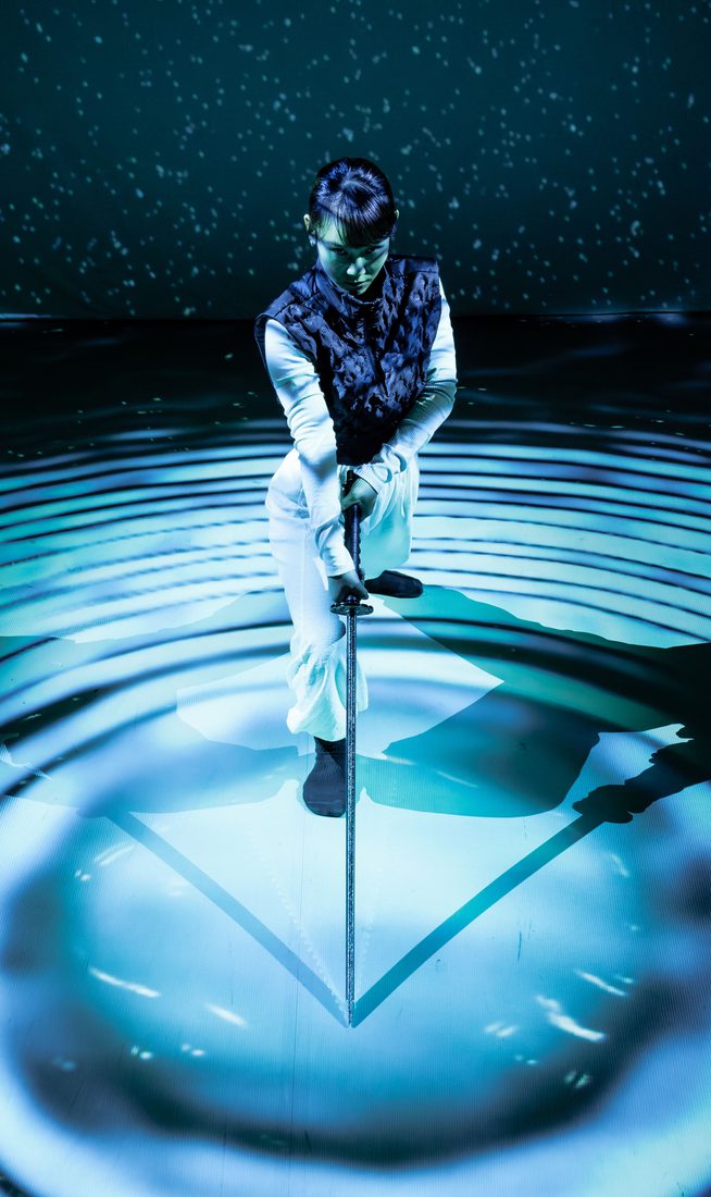 A woman wearing white trousers, a white long-sleeve top, and a navy gilet leans forward onto a long sword, its tip resting at the centre of concentric circles made of blue light and deep shadow. Behind her, a backdrop of scattered light dots against darkness evokes a starry night sky, with a faint green haze resembling northern lights. Her shadow stretches symmetrically to both her left and right, reinforcing the image’s geometric composition. She appears focused and contemplative.