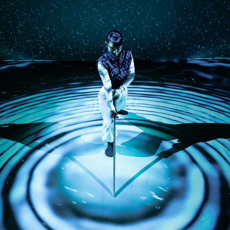 A woman wearing white trousers, a white long-sleeve top, and a navy gilet leans forward onto a long sword, its tip resting at the centre of concentric circles made of blue light and deep shadow. Behind her, a backdrop of scattered light dots against darkness evokes a starry night sky, with a faint green haze resembling northern lights. Her shadow stretches symmetrically to both her left and right, reinforcing the image’s geometric composition. She appears focused and contemplative.