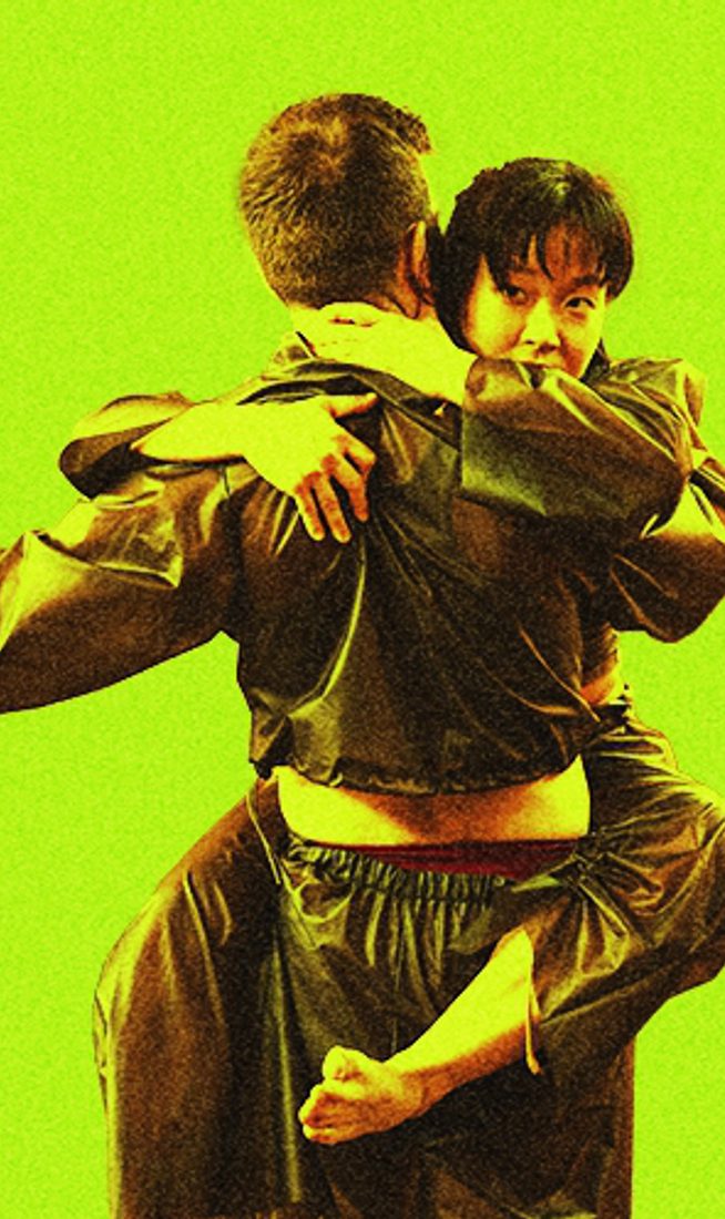 A dancer is shown from the back, wearing a dark, shiny tracksuit, while being embraced by a second dancer whose face is visible over their shoulder. The image is set against a vibrant, solid lime-green background with a slightly grainy, high-contrast texture.